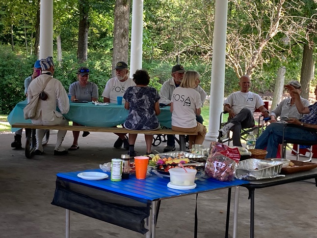 FOSA members seated at a picnic table and chairs with a table of food in the foreground.