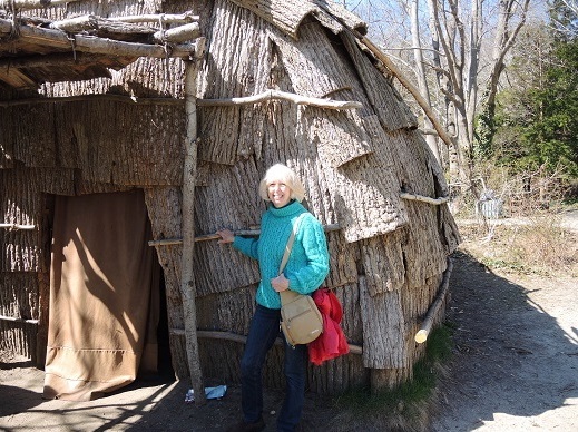 Woman standing in front of building made of tree branches and strips of bark.