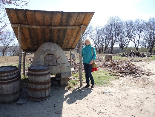 Woman standing beside domed clay oven underneath wooden awning.
