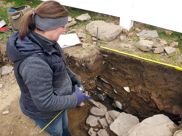 Sarah Sportman bundled up for cold weather in a trench looking at stones and soil profile.